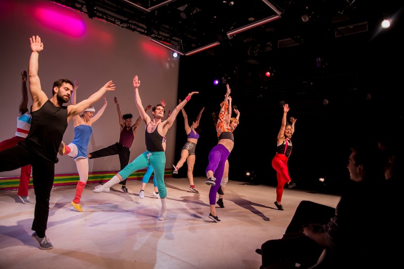 Dancers in colorful 80s style rehearsal clothes with their arms above their heads and right legs extended into the air